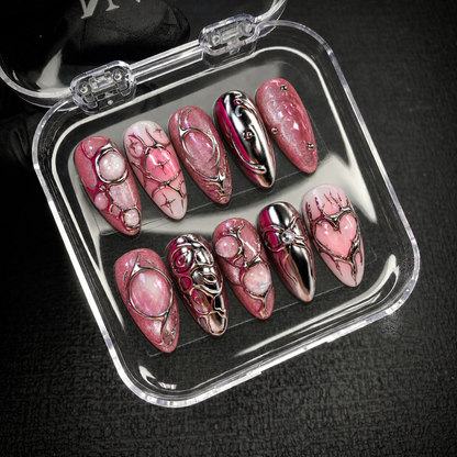 Open clear case holding ten coordinated pink and silver nails on a dark textured surface, with heart, swirl, and opalescent accent details visible.