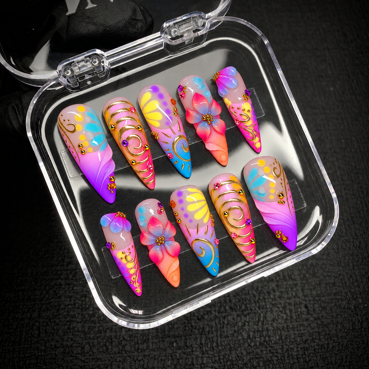 Open clear nail case on a dark textured surface, holding ten coordinated nails with multicolor floral details and gold-colored embellishments.