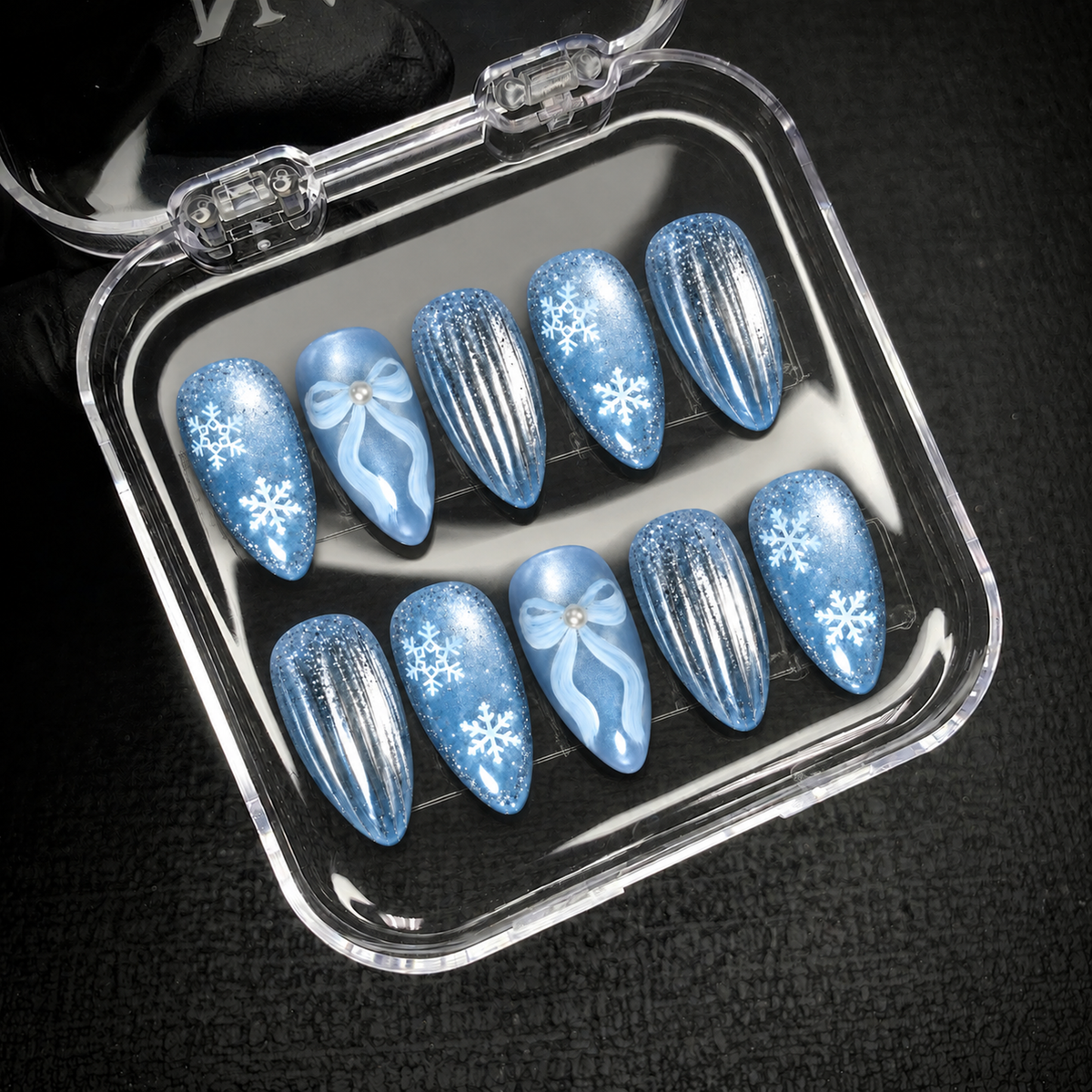 Open clear case holding ten icy blue nails with snowflakes, pearl-like bow centers, and silver stripes on a dark textured surface.