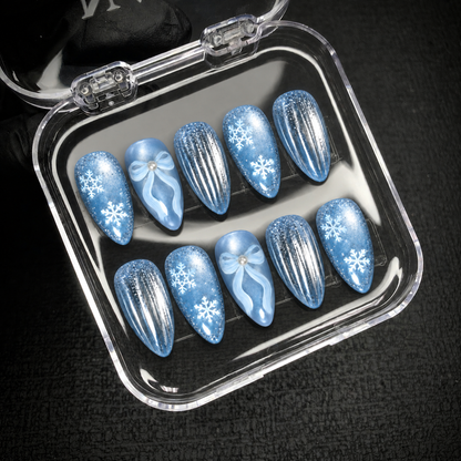 Open clear case holding ten icy blue nails with snowflakes, pearl-like bow centers, and silver stripes on a dark textured surface.