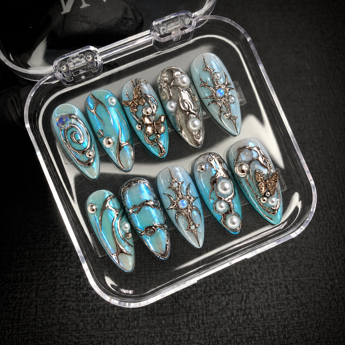 Open clear case holding ten aqua and icy blue nails with silver-tone ornament and butterfly details, photographed at an angle on a dark textured surface.
