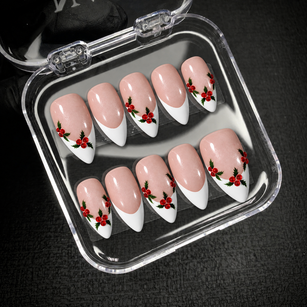 Open clear case on dark textured fabric holding ten coordinated pink shimmer and white French tip nails with holly berry accents.