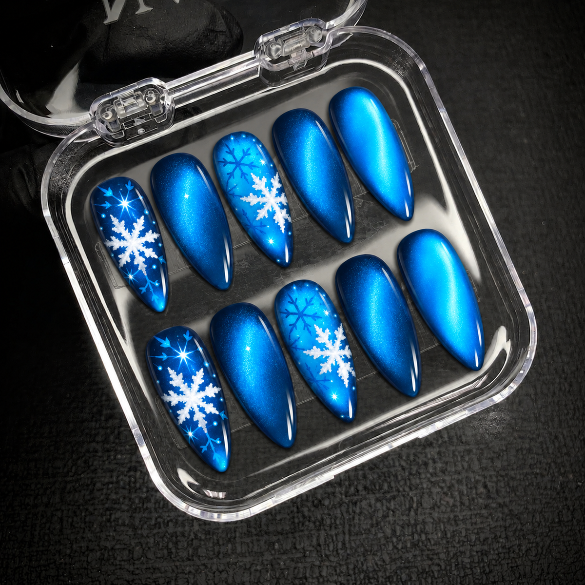 Open clear nail case on a dark textured surface holding ten blue shimmer nails with white and blue snowflake accents.