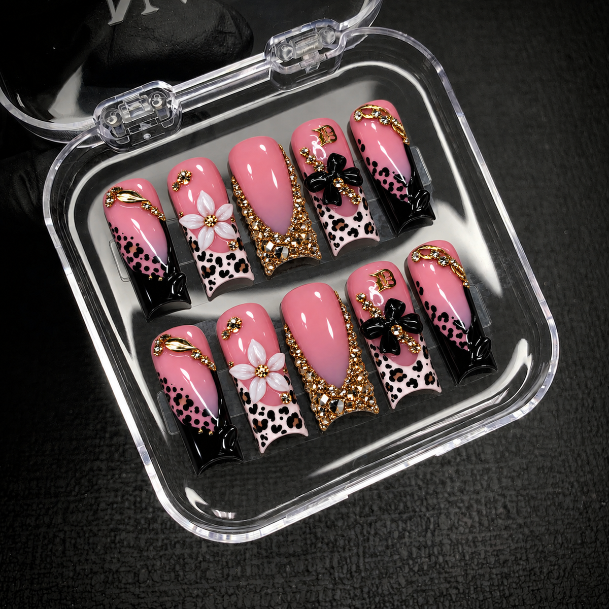 Open clear nail case on dark textured fabric holding ten coordinated pink nails with leopard spots, raised flowers, and gold-tone accents.