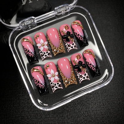 Open clear nail case on dark textured fabric holding ten coordinated pink nails with leopard spots, raised flowers, and gold-tone accents.