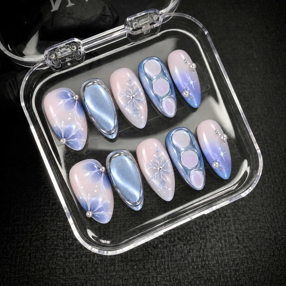 Open clear case holding ten coordinated pink and blue winter nails with snowflakes and pearl accents on a dark textured background.