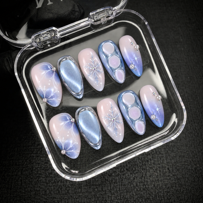 Open clear case holding ten coordinated pink and blue winter nails with snowflakes and pearl accents on a dark textured background.