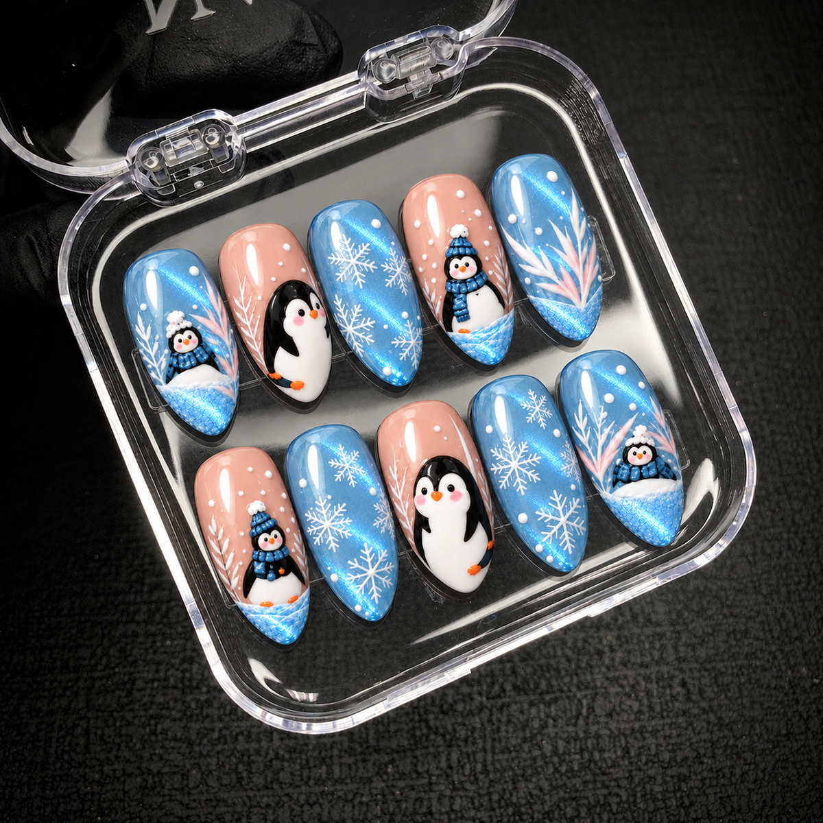 Open clear nail case on a dark textured surface, displaying ten blue and blush pink nails with penguins, white snowflakes, and branch accents.