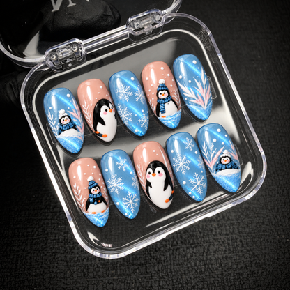Open clear nail case on a dark textured surface, displaying ten blue and blush pink nails with penguins, white snowflakes, and branch accents.