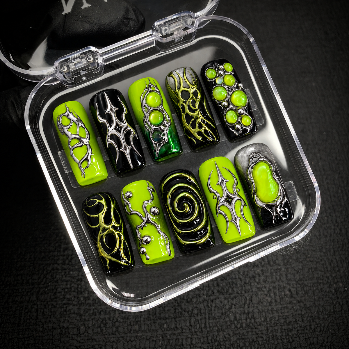 Open clear case holding ten coordinated lime and black nails with metallic-looking details, arranged in two rows on a dark textured surface.
