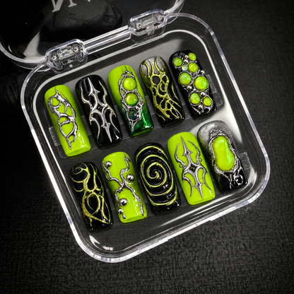 Open clear case holding ten coordinated lime and black nails with metallic-looking details, arranged in two rows on a dark textured surface.
