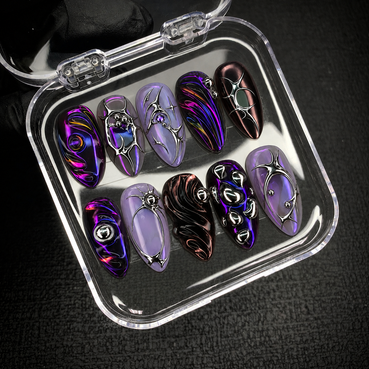 Open clear nail case on a dark textured surface holding ten coordinated violet and lavender nails with reflective silver-tone details.