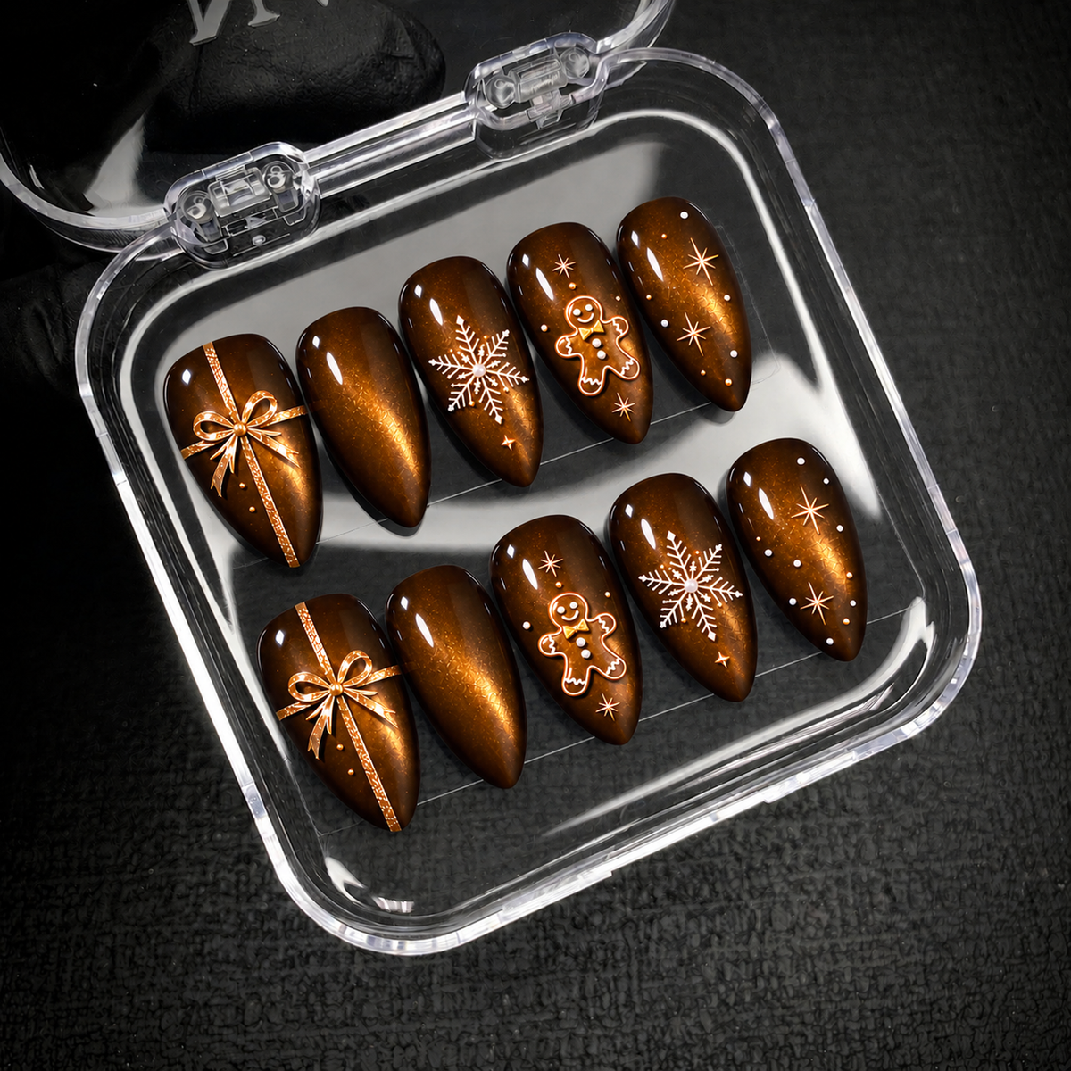 Open clear case holding ten brown and amber Christmas nails with bow, snowflake, gingerbread, and star motifs against a dark textured background.