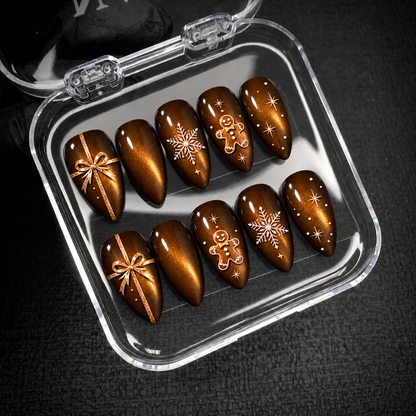Open clear case holding ten brown and amber Christmas nails with bow, snowflake, gingerbread, and star motifs against a dark textured background.