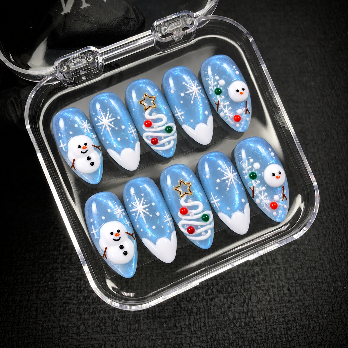 Open clear nail case on dark textured fabric holding ten icy blue nails with snowmen, snowflakes, and swirl Christmas tree designs.