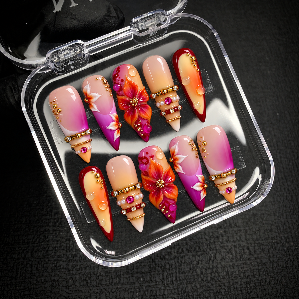 Open clear case displaying ten floral and jewel-accented nails in two rows on a dark textured surface.