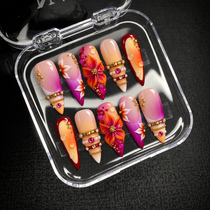 Open clear case displaying ten floral and jewel-accented nails in two rows on a dark textured surface.