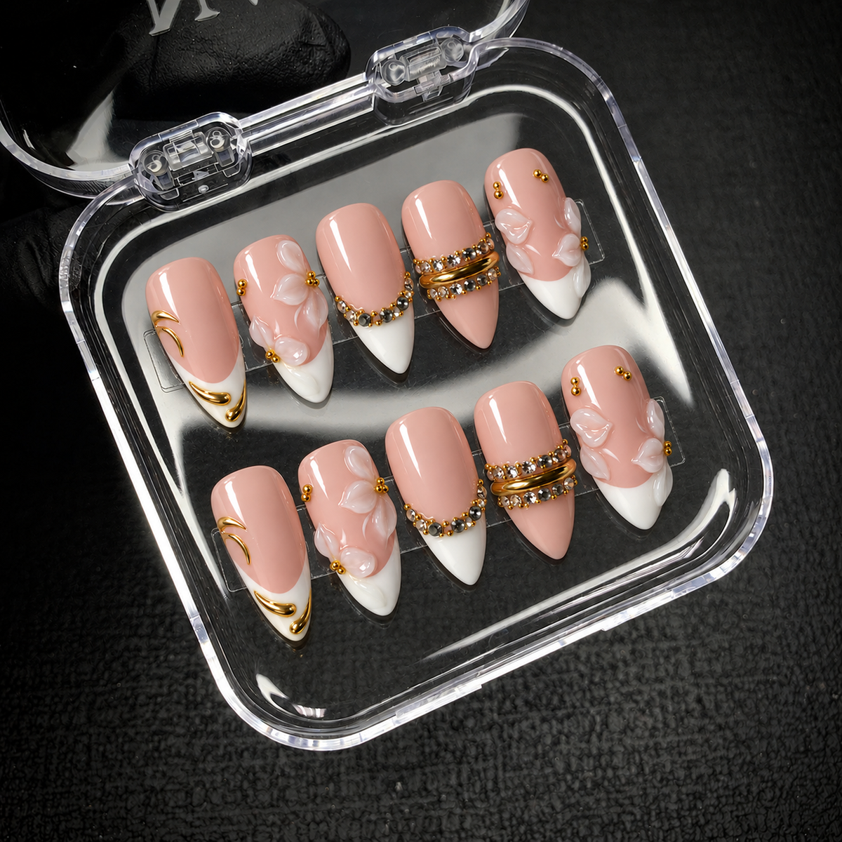 Open clear nail case holding ten blush and ivory nails with raised petals, gold-tone details, and stone accents on a dark textured surface.