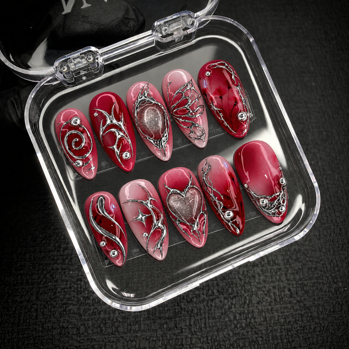 Open clear nail case on dark textured fabric holding ten crimson and blush nails with silver-tone swirls, butterfly-wing details, and shimmering centers.