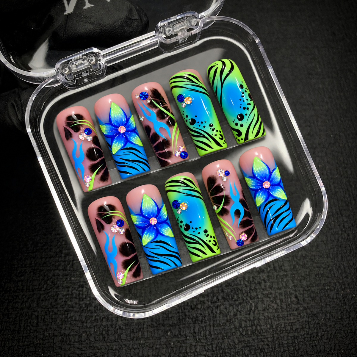 Open clear case holding ten coordinated floral, flame, and animal-inspired patterned nails on a dark textured background.