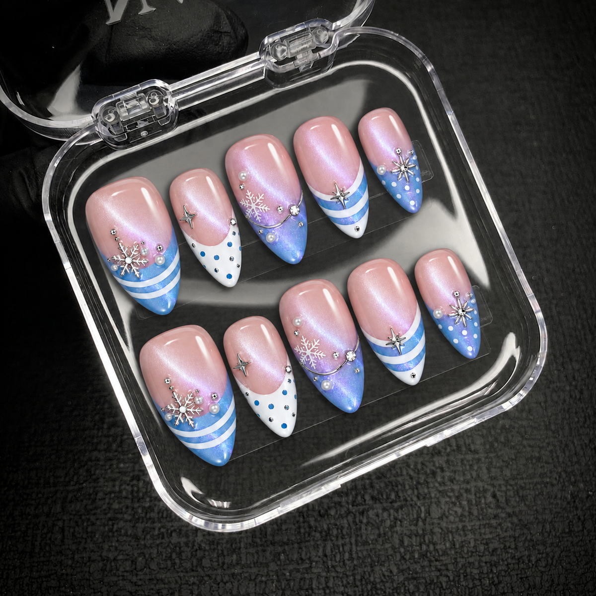 Open clear nail case on dark textured fabric displaying ten pink and blue shimmer nails with snowflakes, stripes, dots, and raised accents.