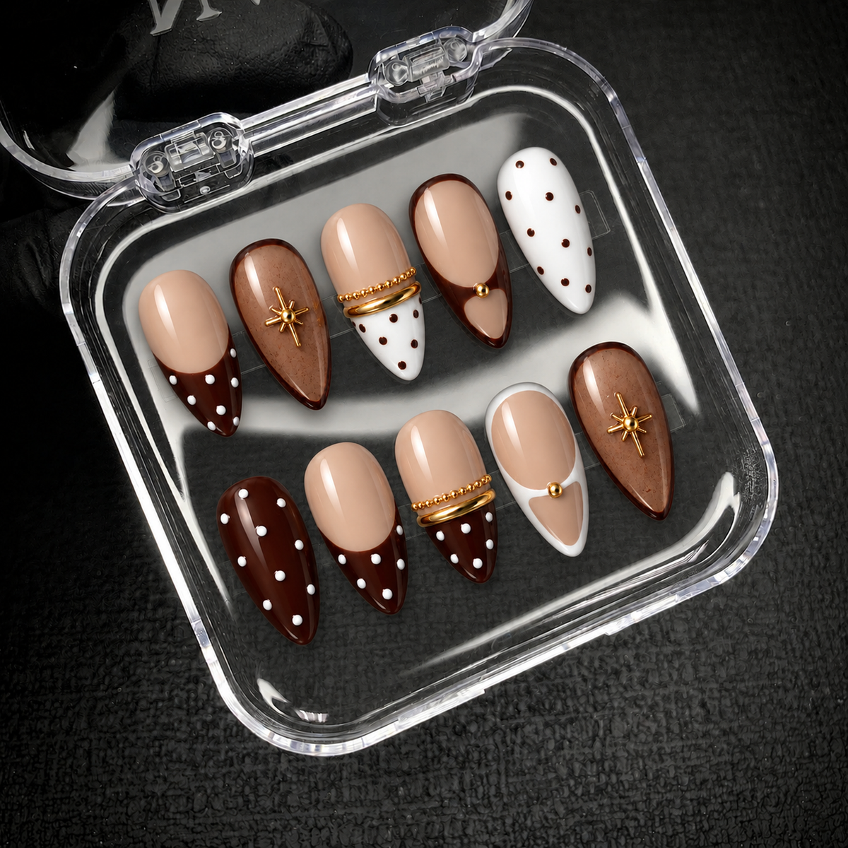Open clear nail case on a dark textured surface holding ten coordinated cocoa, nude, and cream nails with dots and gold accents.