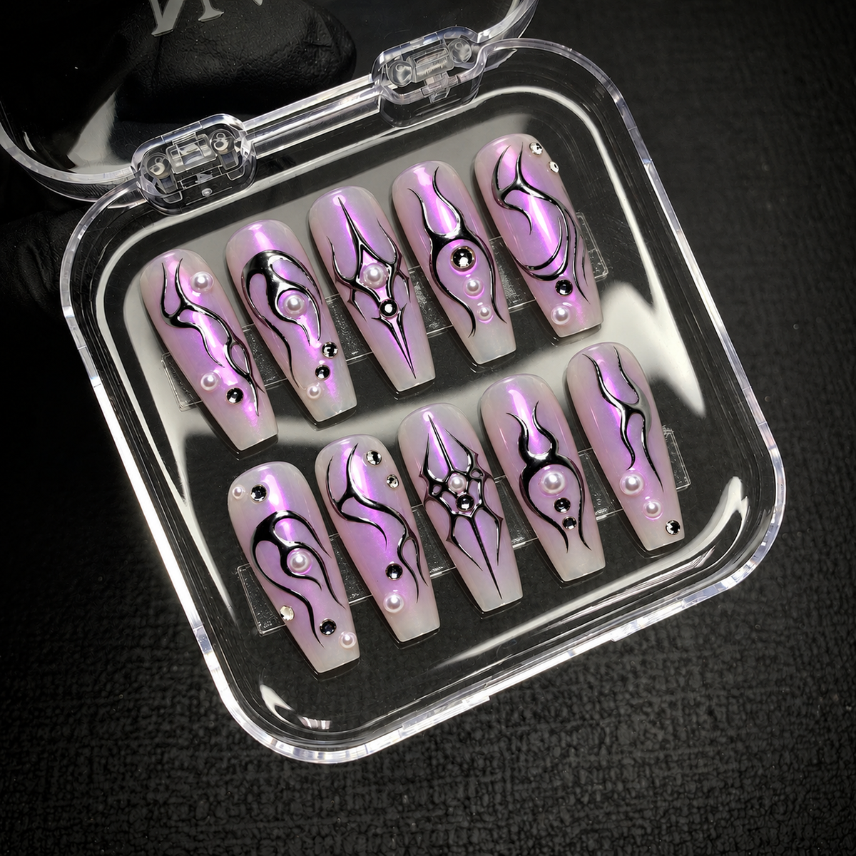 Open clear nail case on a dark textured surface holding ten lilac nails decorated with black sigil motifs, pearl-like accents, and reflective gems.