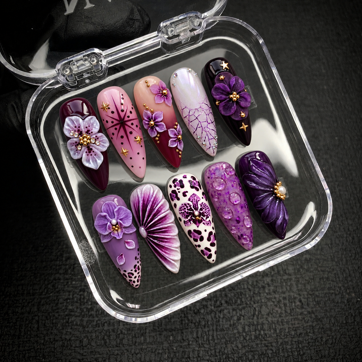 Open clear case holding ten coordinated purple floral nails with gold-tone accents, spotted patterns, and raised petal details on a dark textured surface.