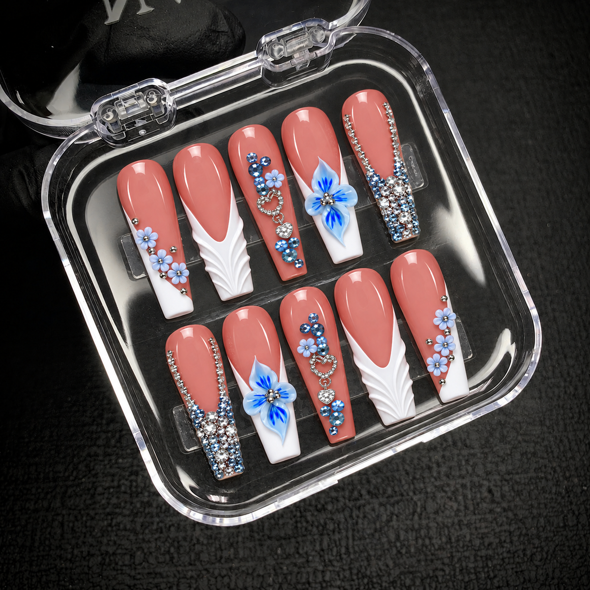 Open clear nail case on dark textured fabric displaying ten coral-pink nails with blue floral details, sculpted white accents, hearts, and rhinestone-covered tips.