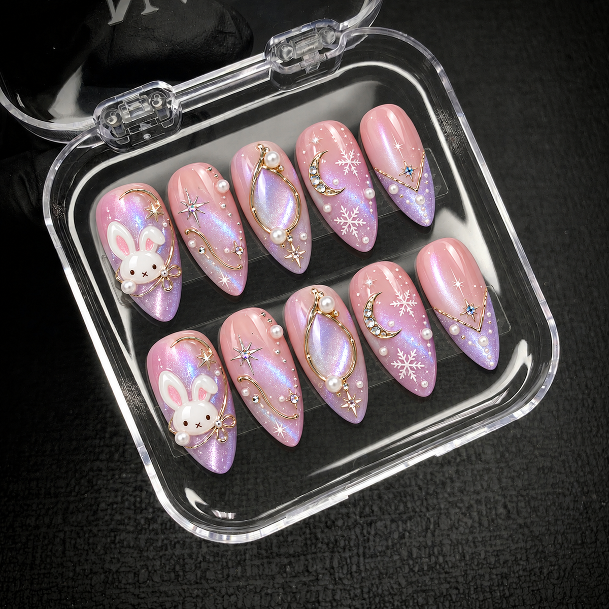 Open clear case on a dark textured surface holding ten coordinated pink and lilac nails with bunny, moon, star, and snowflake designs.
