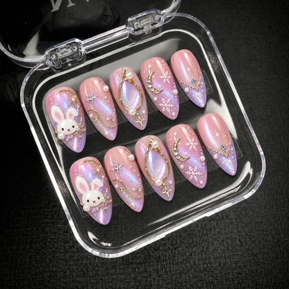 Open clear case on a dark textured surface holding ten coordinated pink and lilac nails with bunny, moon, star, and snowflake designs.