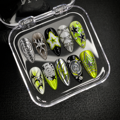 Open clear case on a dark textured surface holding ten coordinated nails with lime accents, silver-toned spirals, black stars, and bow detailing.