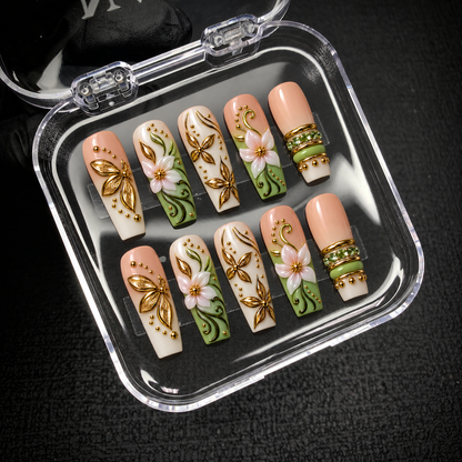 Open clear case displaying ten floral nails on a dark textured surface, with pale blossoms, gold-tone leaves, sage green accents, and decorative bands.