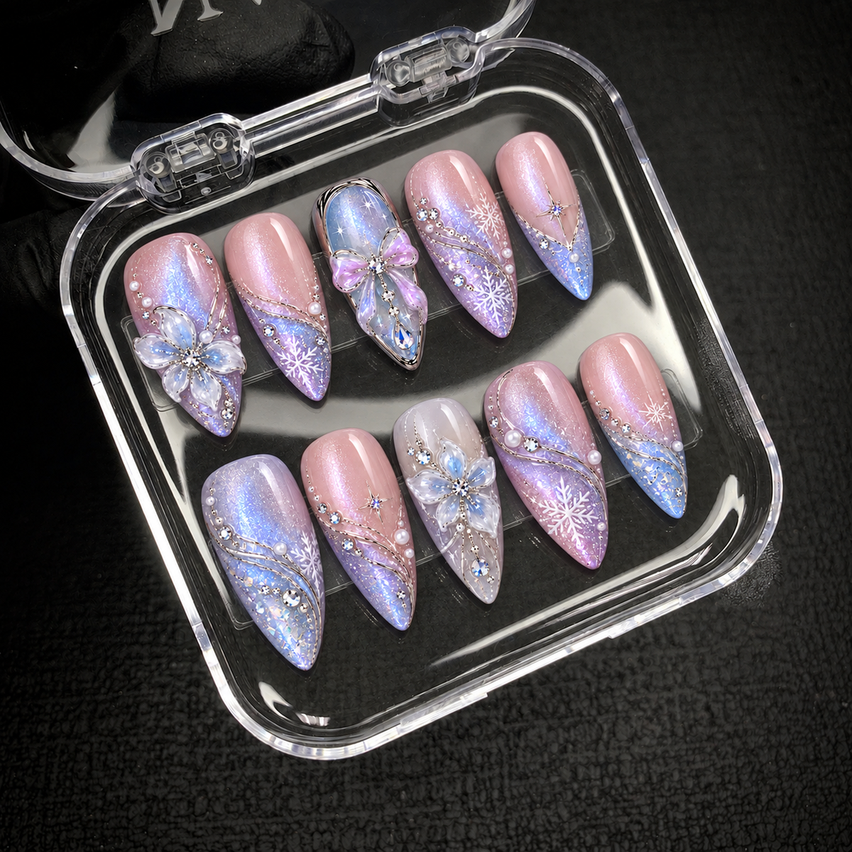 Open clear nail case on a dark textured surface holding ten coordinated pastel nails with snowflakes, silver-toned curves, flower accents, and a bow.