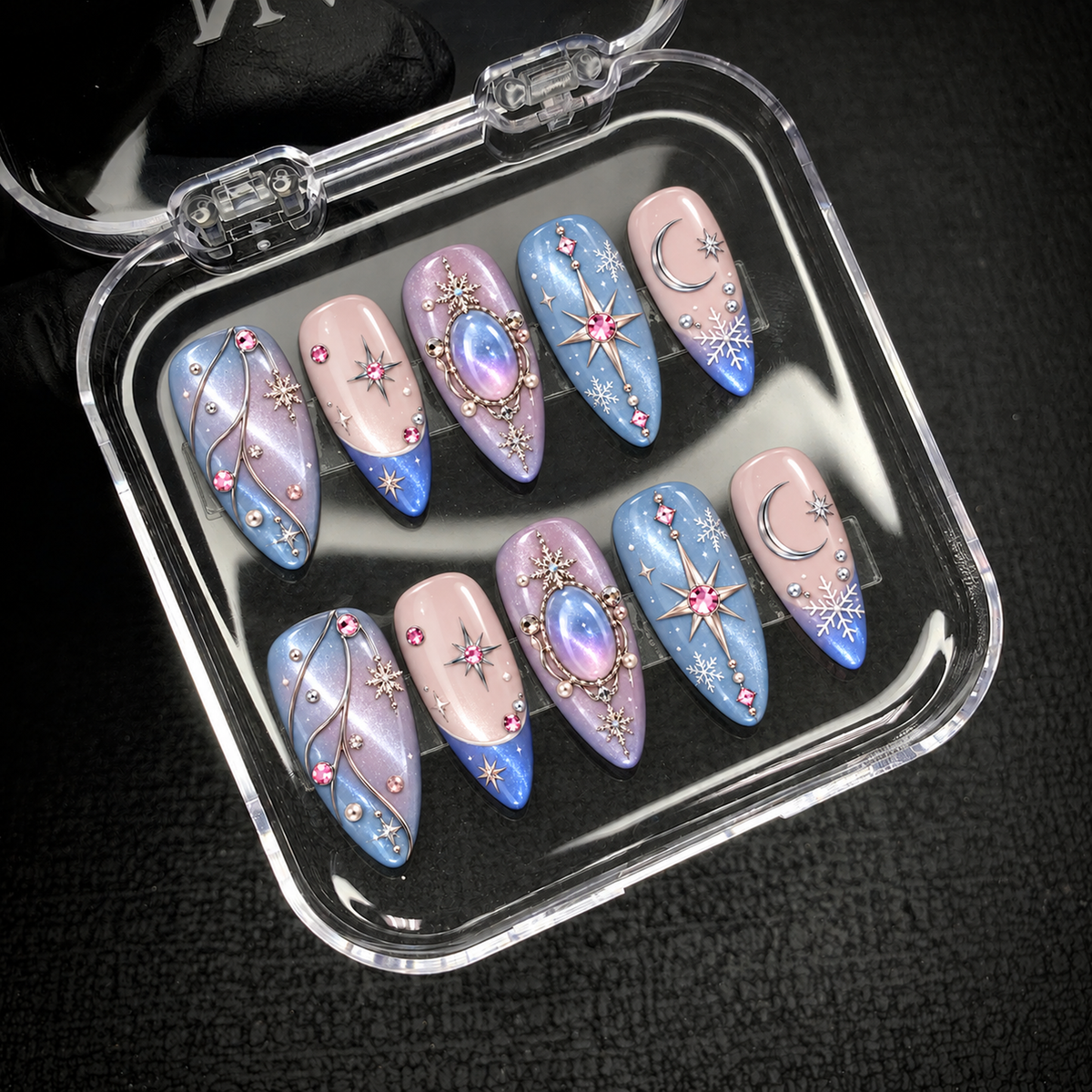 Open clear presentation box holding ten coordinated pastel nails with crescent moons, snowflakes, starbursts, and pink jewel-like accents against a dark textured background.