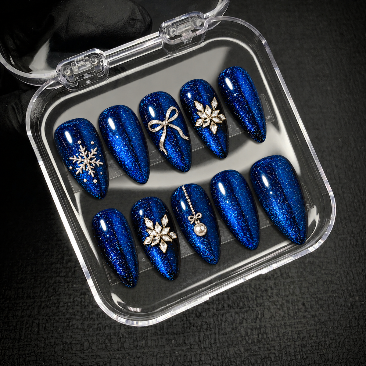 Open clear case holding ten blue shimmer nails with silver-toned winter accents, photographed at an angle on dark textured fabric.