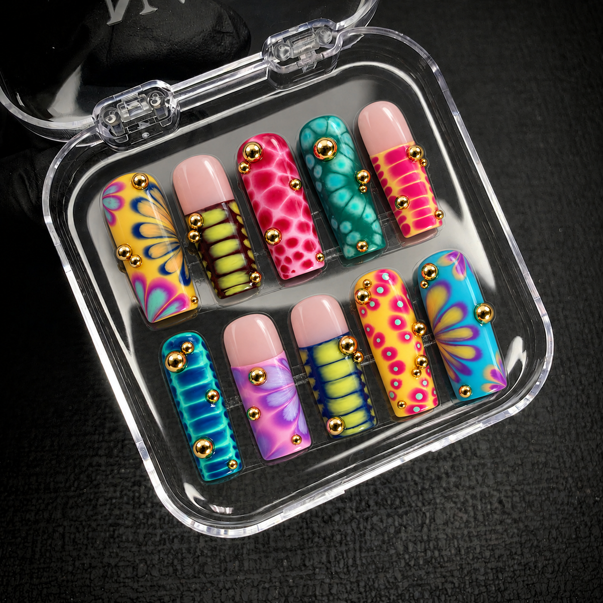 Open clear nail case on a dark textured surface holding ten colorful patterned nails with floral fans and raised gold-tone beads.