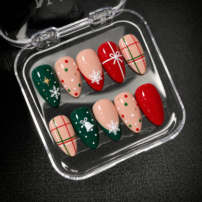 Open clear case on a dark textured surface holding ten coordinated red, evergreen, and peachy nude Christmas nails with white seasonal details.