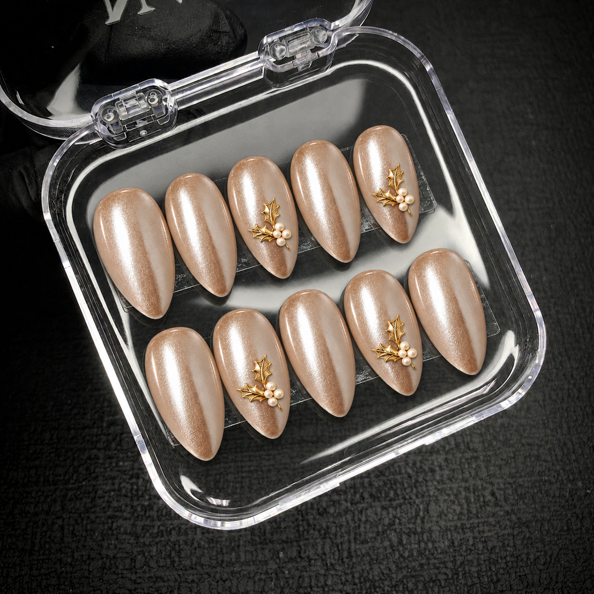 Open clear nail case on a dark textured surface holding ten champagne shimmer nails, including four with holly leaf and pearl-like accents.