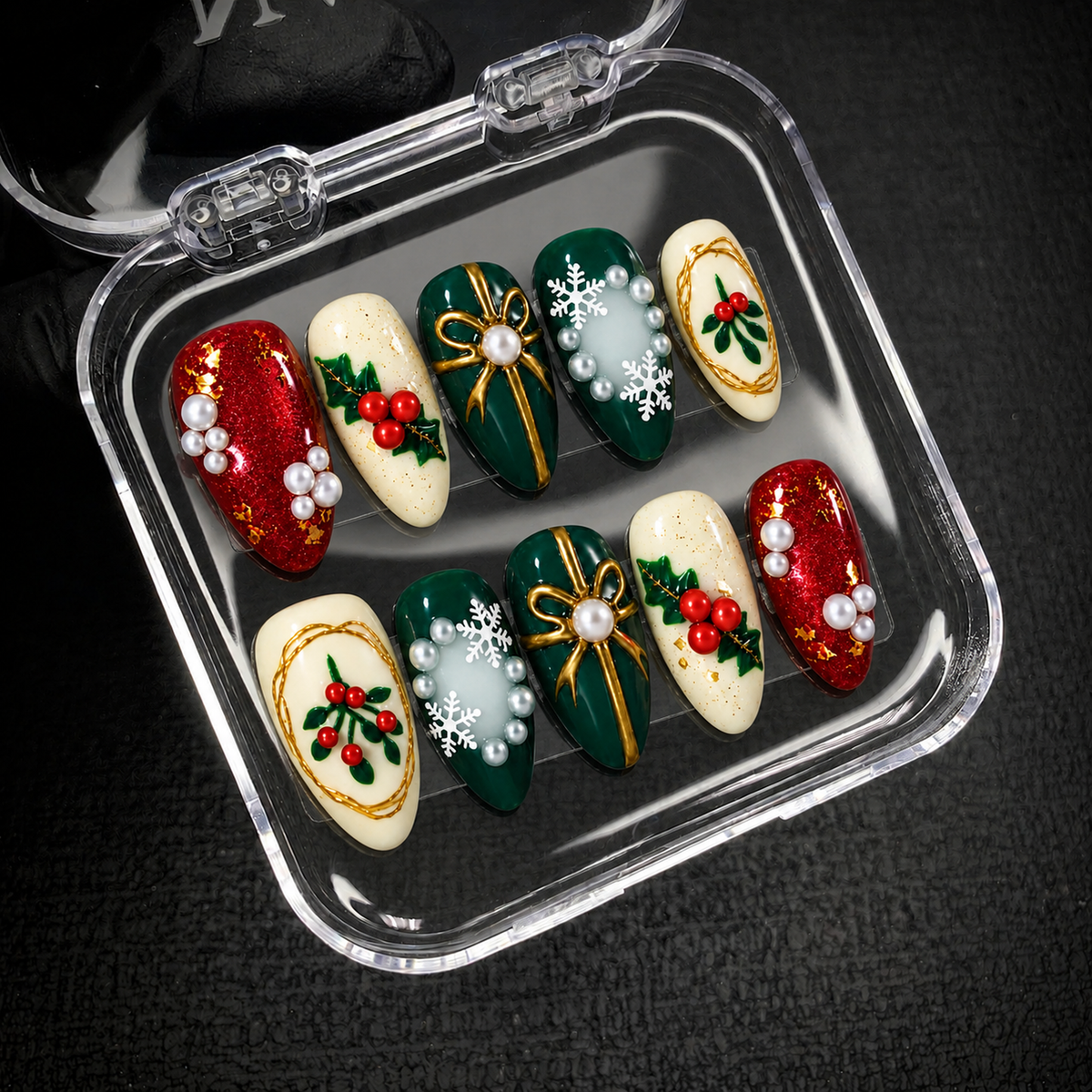 Open clear nail case on a dark textured surface holding ten coordinated Christmas nails with gold-tone bows, berry sprigs, snowflakes, and pearl-like embellishments.