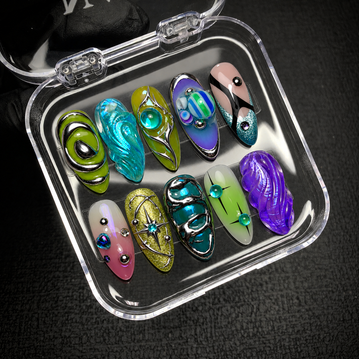 Open clear nail case on a dark textured surface holding ten coordinated nails with colorful waves, silver-colored outlines, and orbital details.