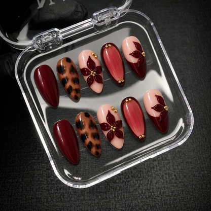 Open clear nail case holding ten coordinated burgundy, leopard-print, and blush floral nails on a dark textured background.