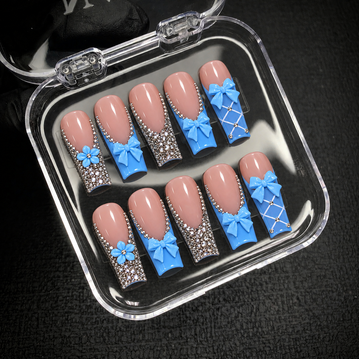 Open clear nail case on a dark textured surface holding ten coordinated pink and blue nails with bows, flowers, and rhinestone details.