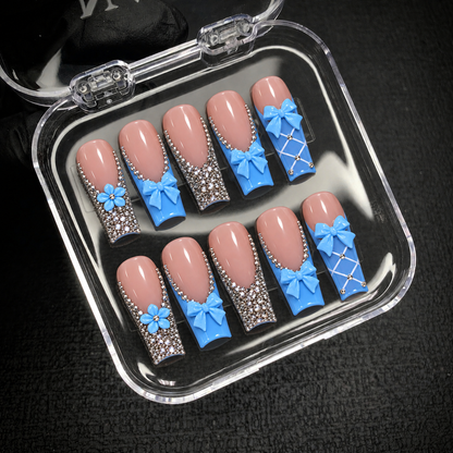 Open clear nail case on a dark textured surface holding ten coordinated pink and blue nails with bows, flowers, and rhinestone details.