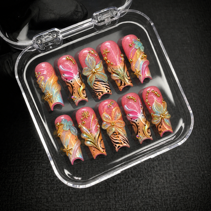 Open clear case holding ten coordinated coral-pink nails with raised flowers, flowing swirls, black patterns, and gold-toned starfish accents on dark fabric.