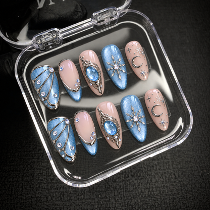 Moonlit Frost nails arranged in two rows inside an open clear case, with blue shimmer, blush pink, snowflakes, a crescent moon, and silver-tone accents visible.