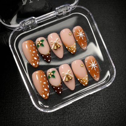 Open clear case holding ten Christmas press-on nails on dark textured fabric, showing holly, bow and bell accents, and white snowflakes.