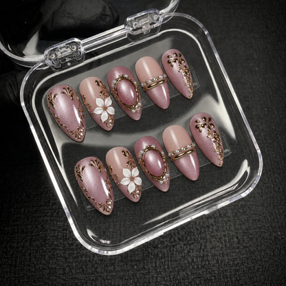 Open clear case on dark textured fabric displays ten mauve and blush nude nails with gold-toned flourishes, white flowers, and decorative bands.