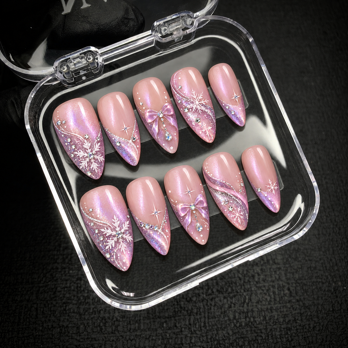 Open clear nail case on dark textured fabric holding ten pink nails decorated with lavender glitter, white snowflakes, dimensional bows, and gems.