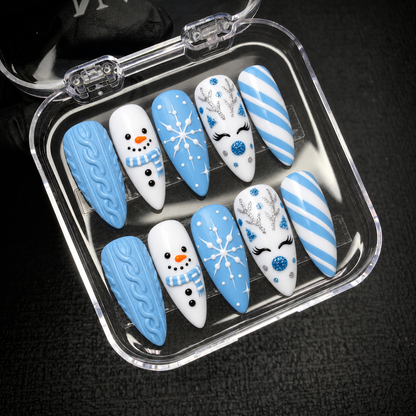 Open clear case on a dark textured background holding ten coordinated blue and white nails with sweater patterns, snowmen, snowflakes, reindeer accents, and stripes.